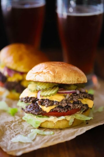 Two best-ever smash burgers with toasted brioche buns sitting on crumpled brown paper with two glasses of dark beer in the background.