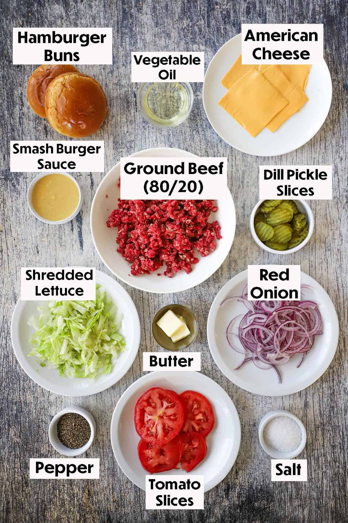 An arrangement of ingredients for best-ever smash burger on a grey wooden background including ground beef, buns, oil, cheese slices, burger sauce, pickles, lettuce, onion, butter, tomato slices, salt, and pepper.