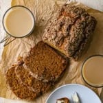 An overhead view of a loaf of best homemade pumpkin bread that has three slices cut from it and resting on brown paper with two glass mugs of creamed coffee nearby.