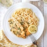 An overhead view of an elegant white dinner plate filled with a serving of homemade chicken piccata with a pile of linguine and a lemon wedge off to the side.