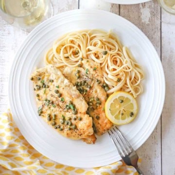 An overhead view of an elegant white dinner plate filled with a serving of homemade chicken piccata with a pile of linguine and a lemon wedge off to the side.