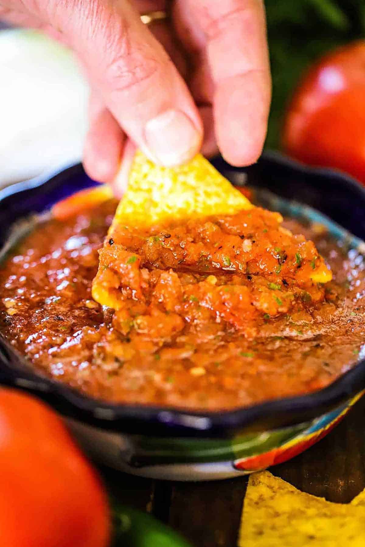 A person raising a triangular corn tortilla chip from a festive bowl filled with roasted tomato salsa.