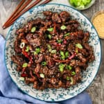 An overhead view of a colorful serving bowl filled with crispy chili beef garnished with slivers of red peppers, chopped scallions, and toasted sesame seeds.