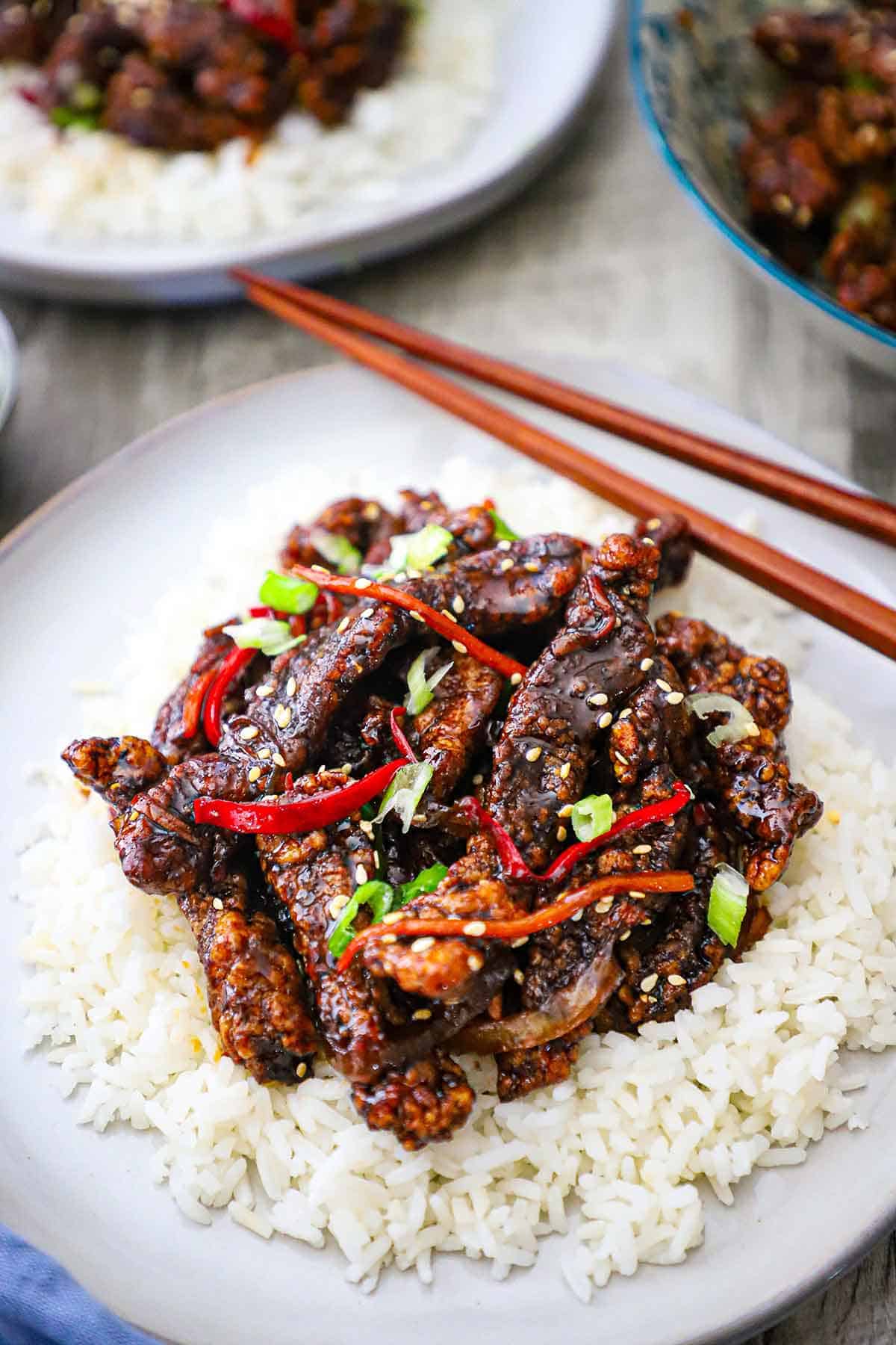 A light grey dinner plate filled with a serving of crispy chili beef on a bed or steamed rice with chop sticks sitting on the edge of the plate.