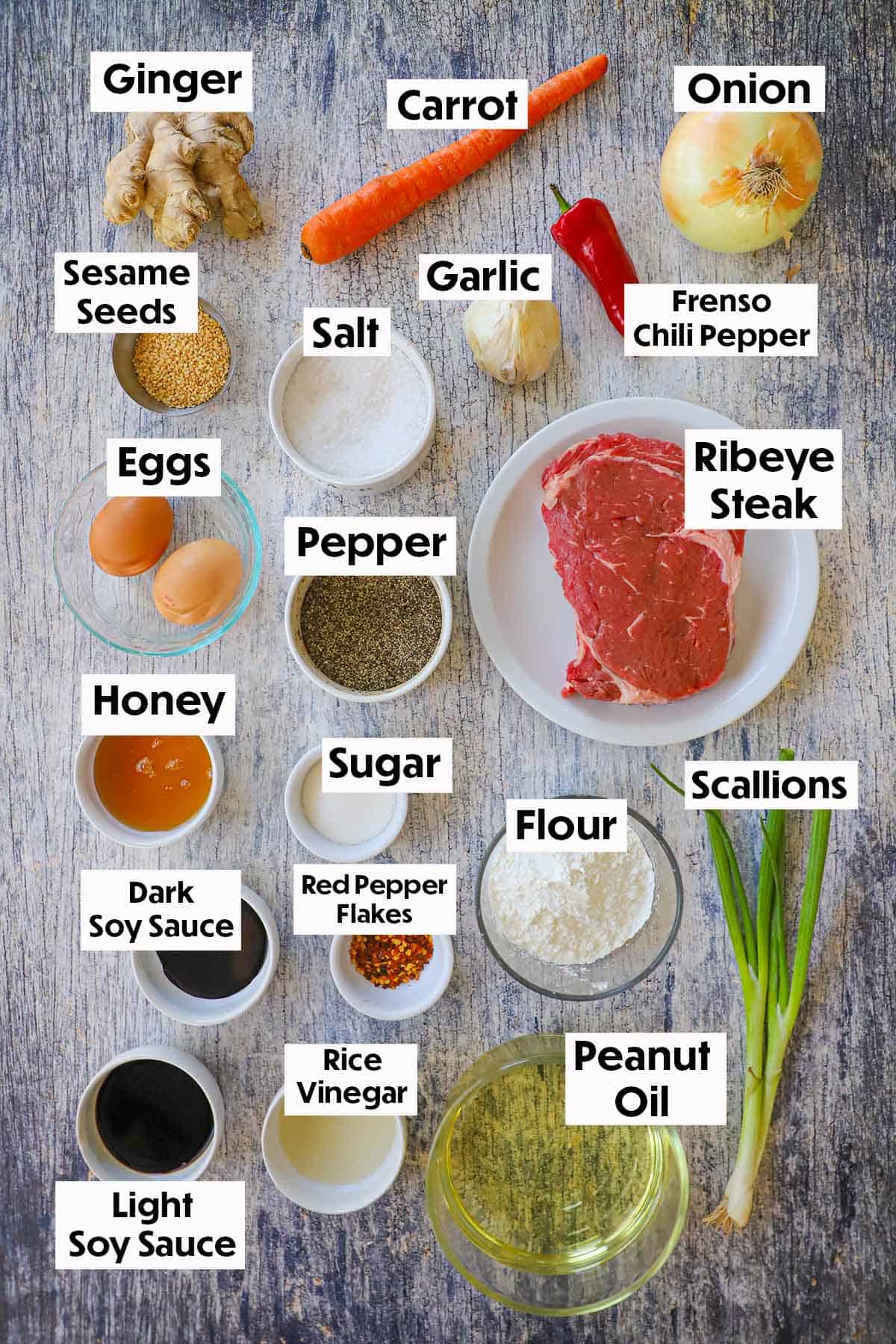 An arrangement of ingredients for crispy chili beef on a grey wooden background including a raw ribeye steak, ginger, carrot, onion, garlic, eggs, soy sauce, flour, oil, scallions, and seasonings.