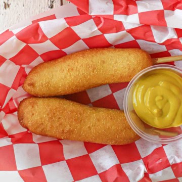 Two homemade corn dogs next to each other with a small container of yellow mustard nearby all on a red and white checkered wax paper in a basket.