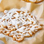 A close-up view of a homemade funnel cake topped with a dusting of powdered sugar and is resting on a brown piece of paper.