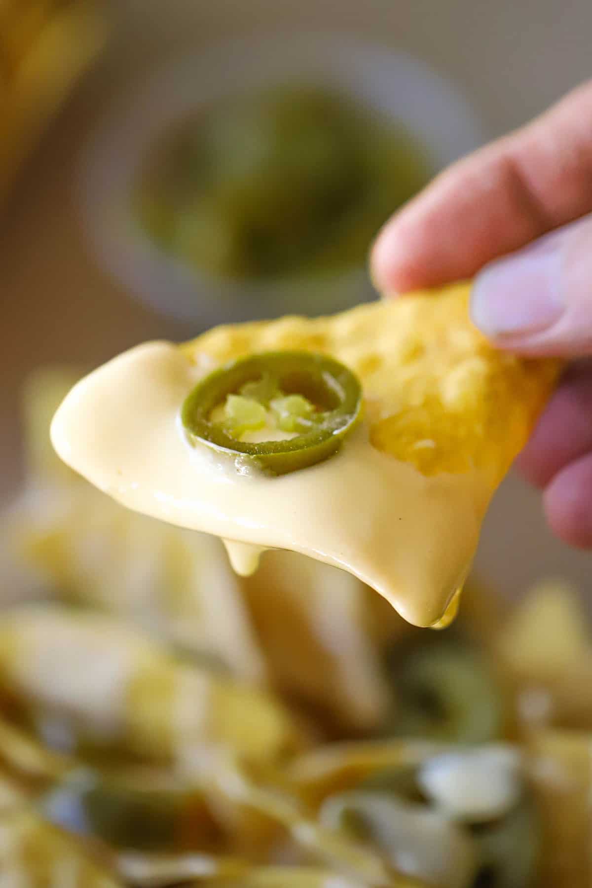 A person holding a corn tortilla chip that is partially coated with easy homemade nacho cheese sauce and topped with a single pickled jalapeño slice.