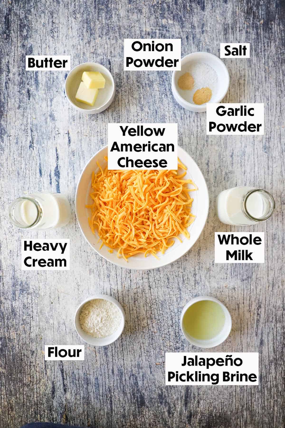 An arrangement of ingredients for easy homemade nacho sauce on grey wooden background including shredded yellow American cheese, butter, seasonings, cream, milk, flour, a jalapeño pickling brine.