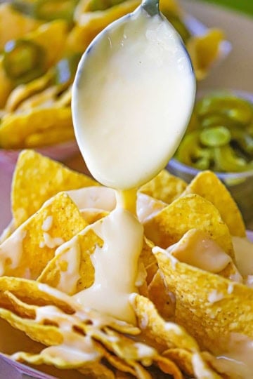 A spoon being used to drizzle easy homemade nacho cheese sauce onto rows of tortilla chips that are sitting side by side in a paper basket.