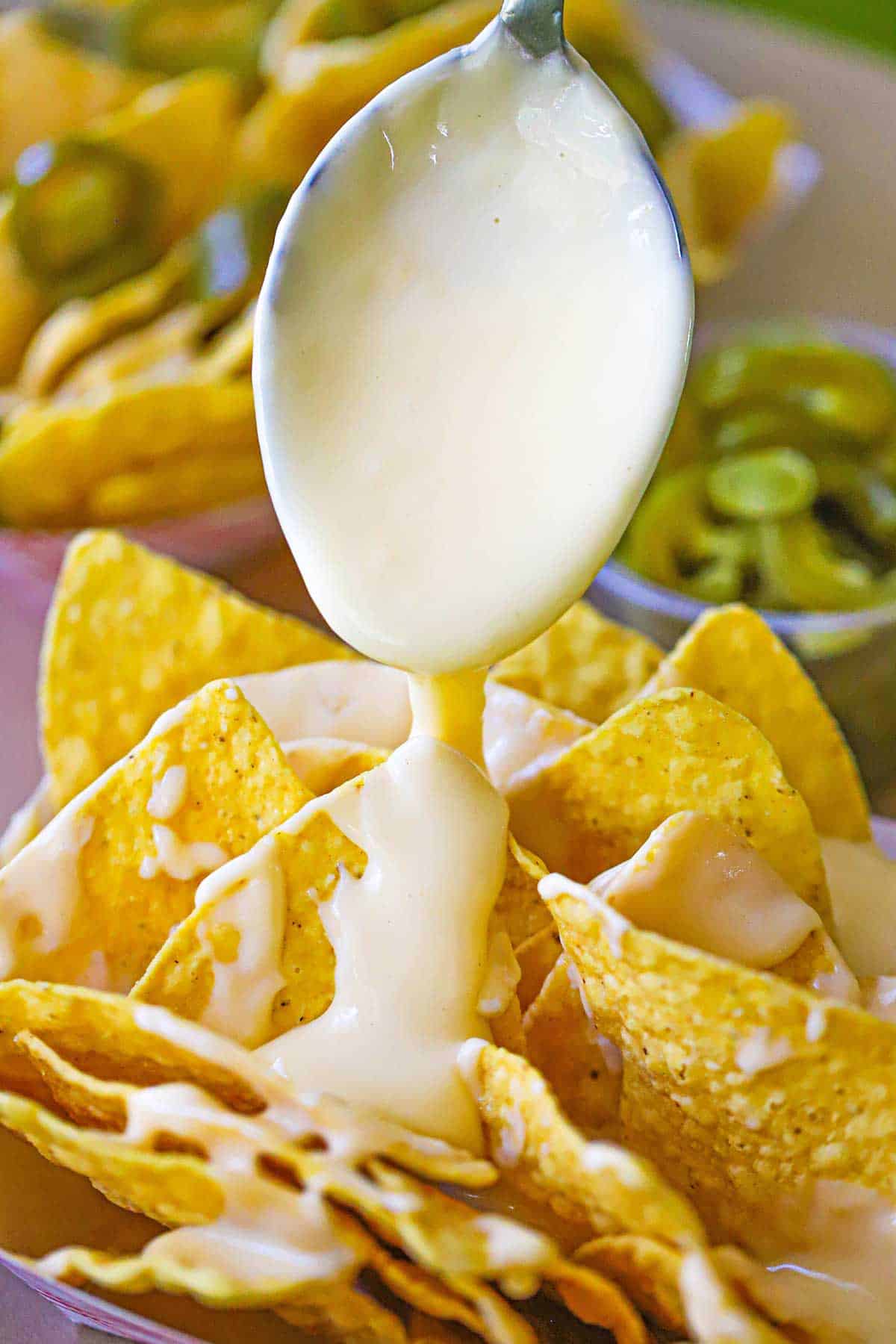 A spoon being used to drizzle easy homemade nacho cheese sauce onto rows of tortilla chips that are sitting side by side in a paper basket.