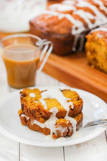 Two slices of pumpkin butterscotch bread on a white plate next to a cup of coffee and two loaves of bread.