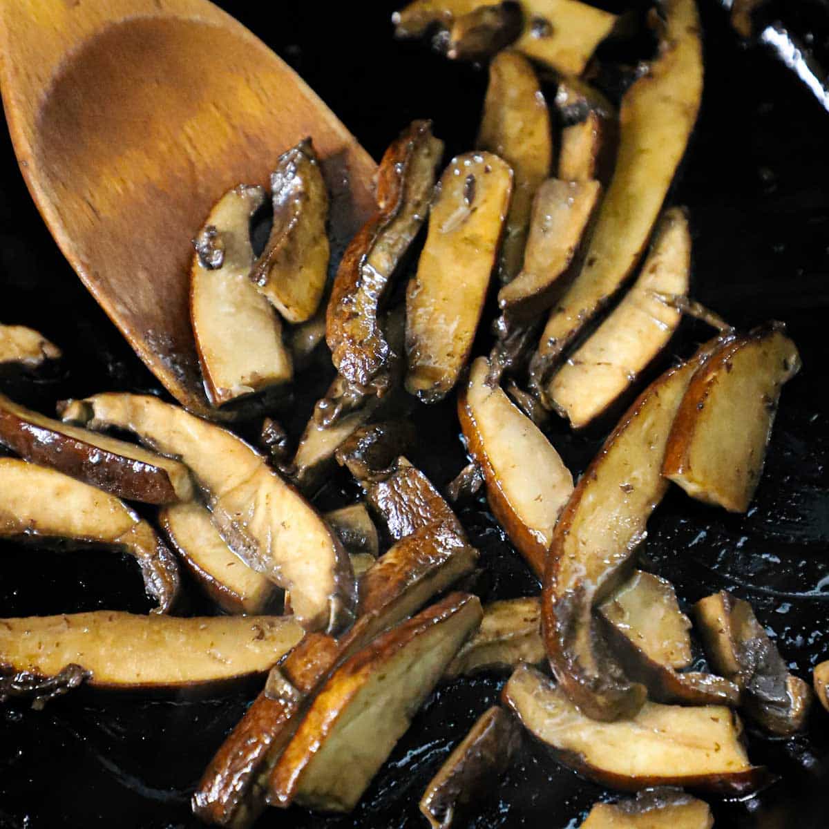 A wooden spoon being used to stir and sauté sliced portobello mushrooms in a cast-iron skillet.