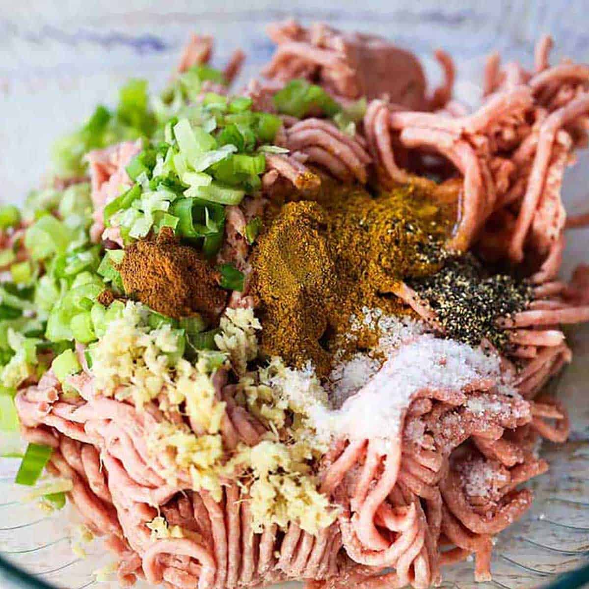A pile of uncooked turkey in a glass bowl topped with curry powder, salt, pepper, chopped ginger, garlic, and scallions.