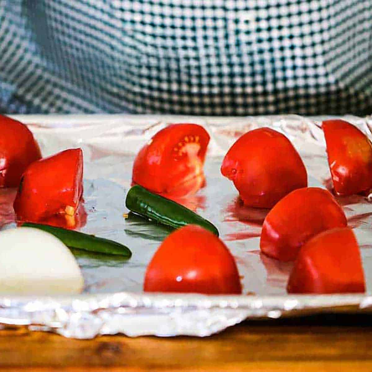 A person holding an aluminum foil-lined baking sheet the is holding tomatoes that have been quartered, two serrano peppers, and a quarter of a white onion.
