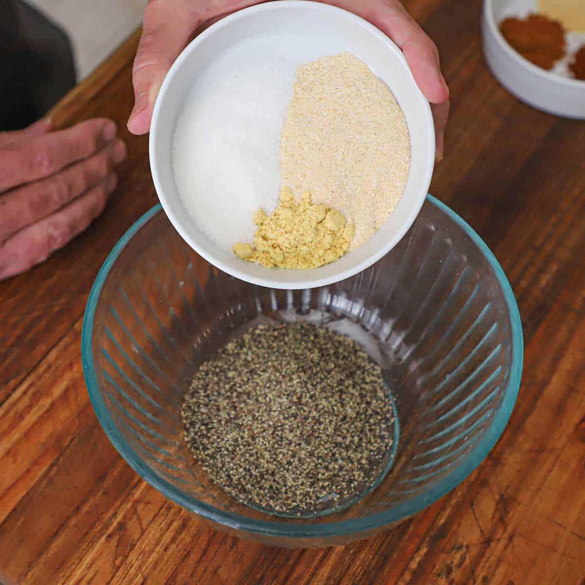 A person dumping onion powder, sugar, and ground mustard into a glass bowl that has ground pepper in it.