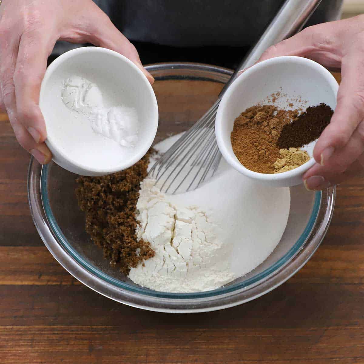 A person holding a small white bowl in one hand that contains small mounds of salt, baking soda, and baking powder and another white bowl in his other hand containing pumpkin bread spices over a bowl of flour and sugar.