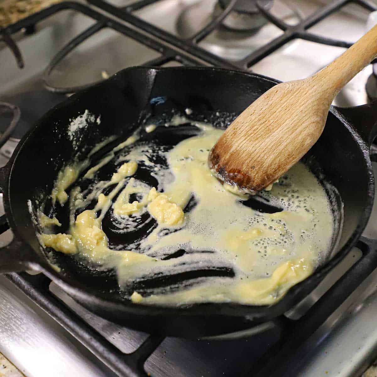 A person using a wooden spatula to stir a butter and flour roux in a black cast-iron skillet on a gas stove.