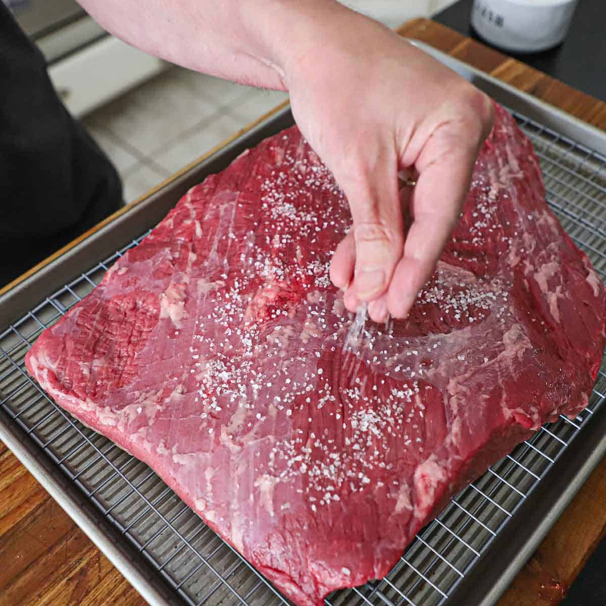 A person using his fingers to sprinkle coarse Kosher salt all over the surface of an uncooked brisket flat that is resting on baking rack inside a baking sheet.