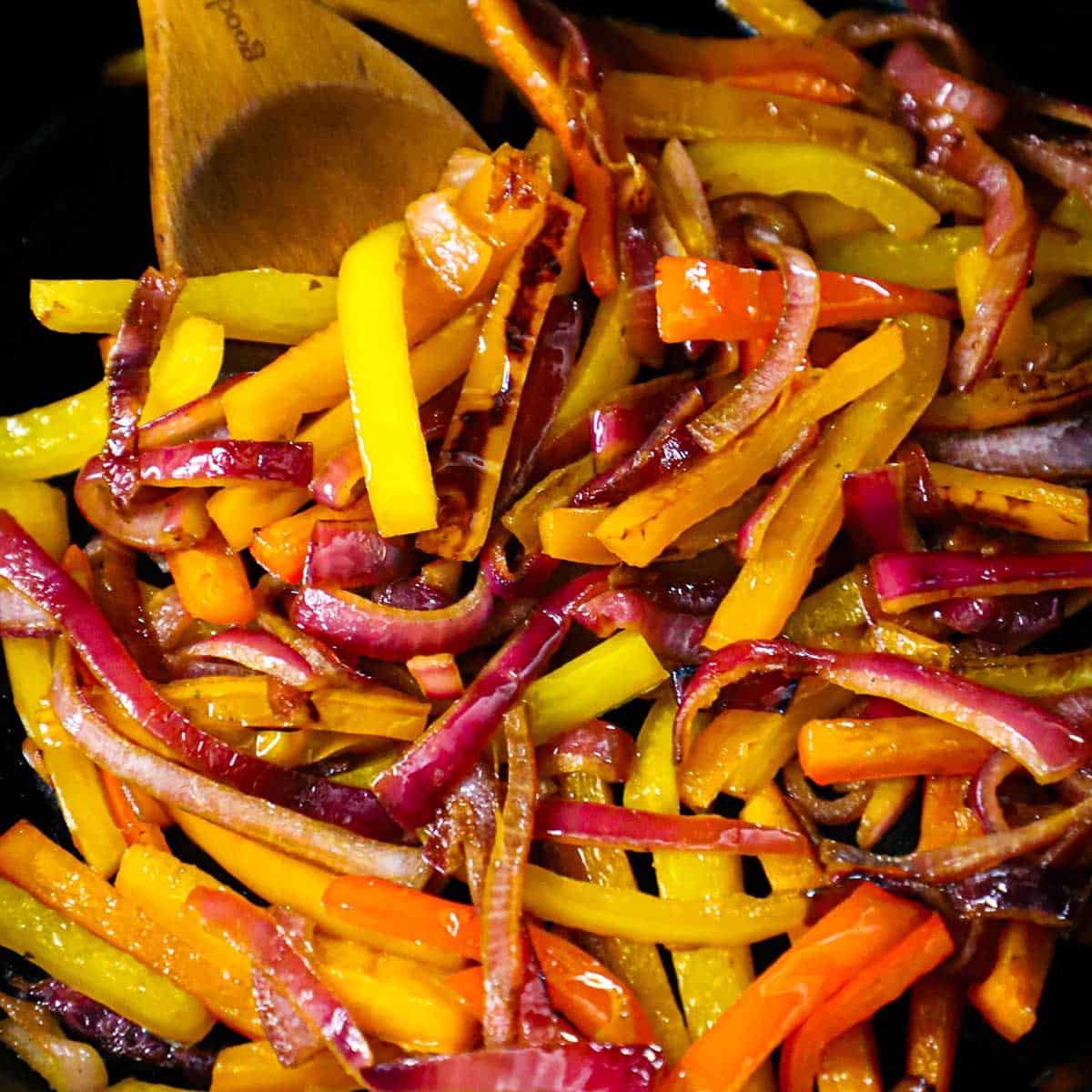 A wooden spoon being used to stir and sauté strips of red, yellow, and orange bell peppers, as well as caramelized onions.