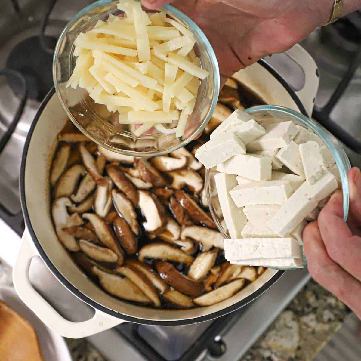 A person holding a small glass bowl filled with strips of bamboo shoots in one hand and the other hand holding a small glass bowl filled with strips of firm tofu both over a pot filled with a simmering broth with sliced mushrooms floating at the surface.
