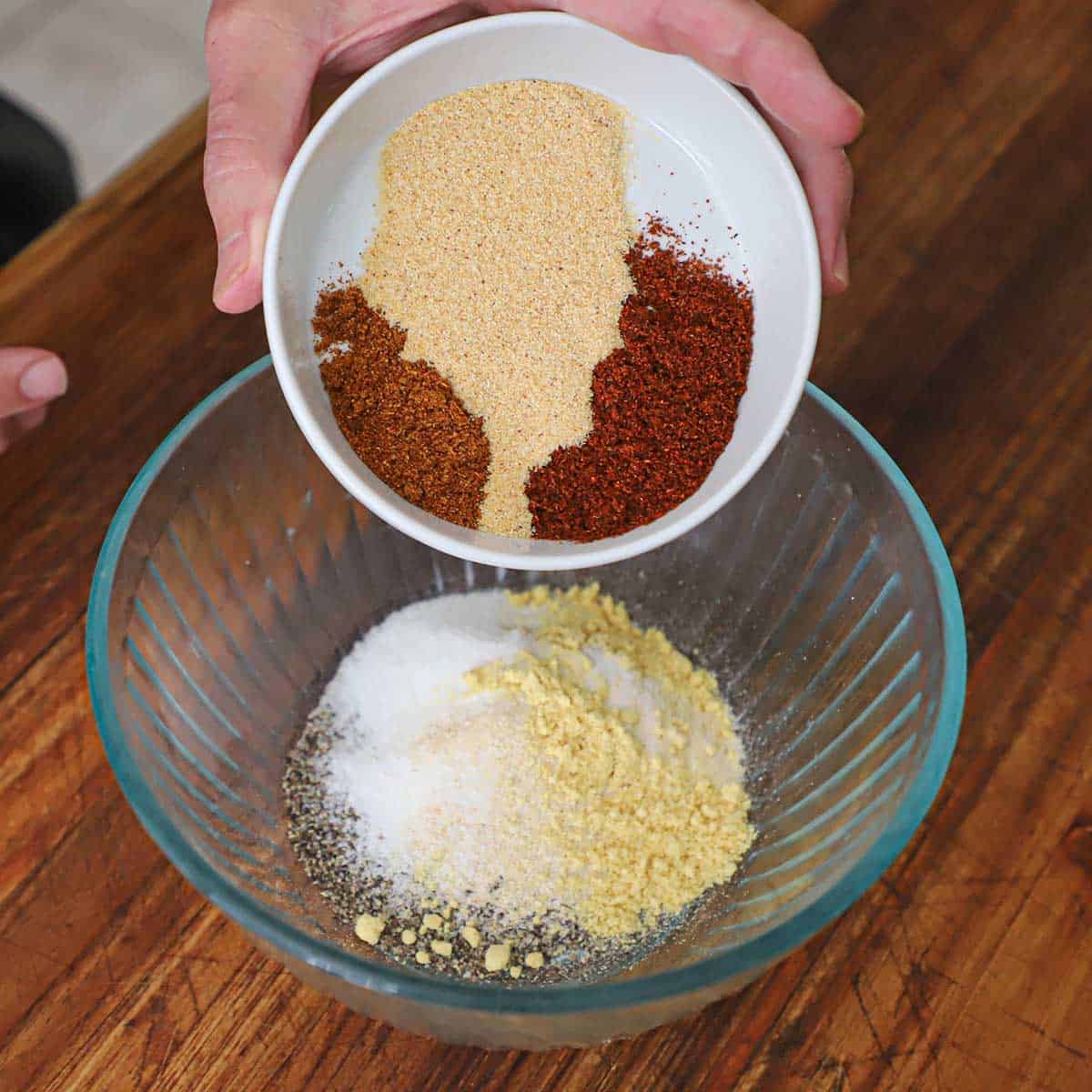 A person dumping small piles of cayenne pepper, garlic powder, and chili powder from a small white bowl that is filled with piles of black pepper, sugar, onion powder, and ground mustard.