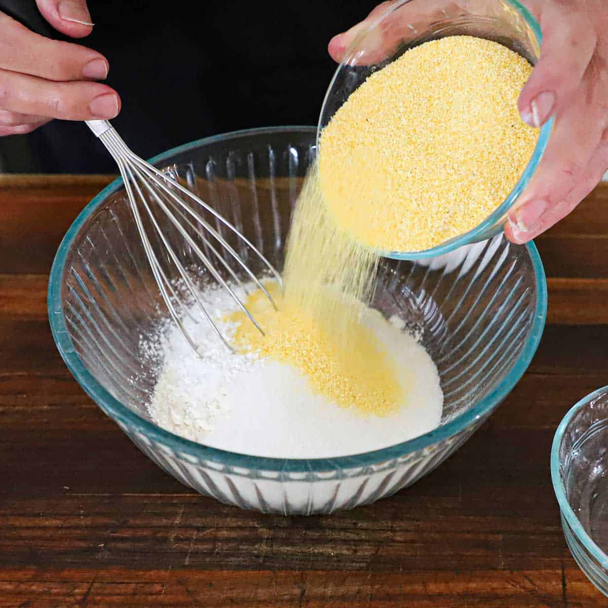 A person pouring cornmeal from a glass bowl into another glass bowl filled with flour, sugar, and baking powder.