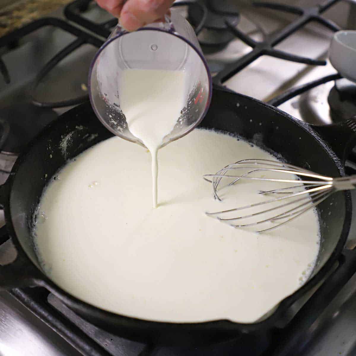 A person pouring cream into a cast-iron skillet that has a roux in it on a gas stove.