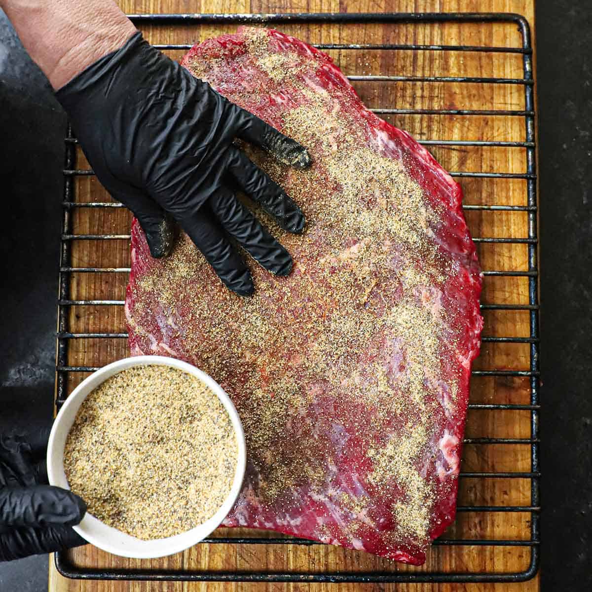 A person who is wearing latex black gloves is pressing a beef run all over the surface of an uncooked brisket flat on the grate of a smoker that is sitting on a wooding cutting board.