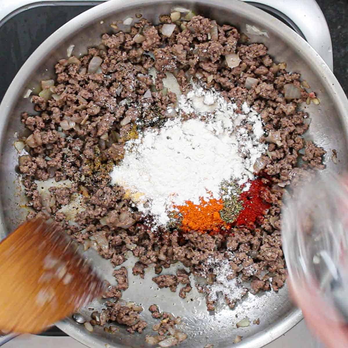 A person adding Tex-Mex seasonings and flour to cooked ground beef in a large skillet on a gas stove.