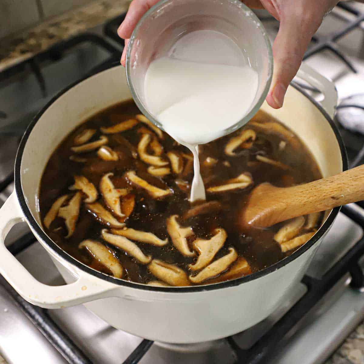 A person pouring a cornstarch slurry from a small glass bowl into a pot of simmering homemade Chinese hot and sour soup.