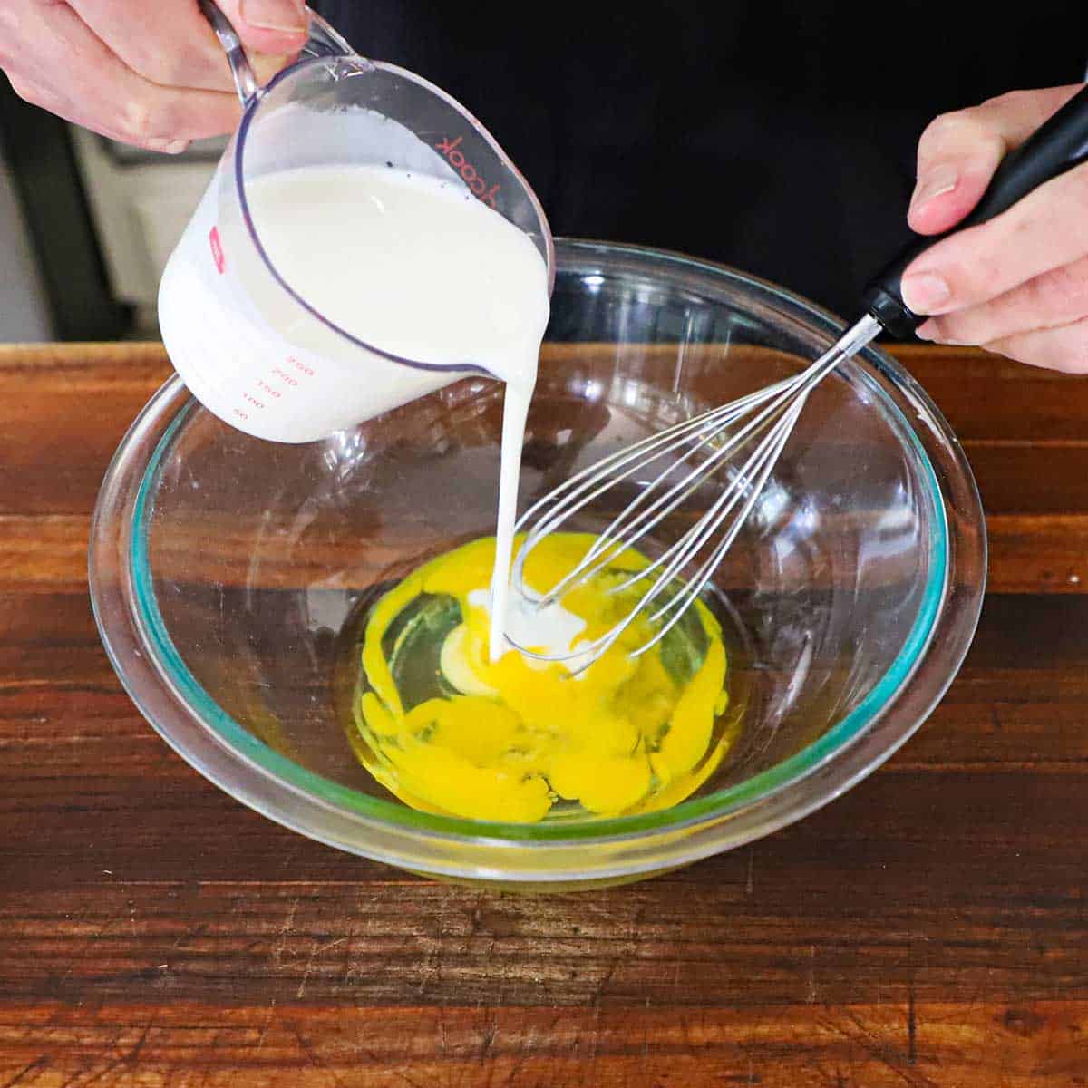 A person pouring buttermilk from a measuring cup into a glass bowl filled with two lightly beaten eggs.