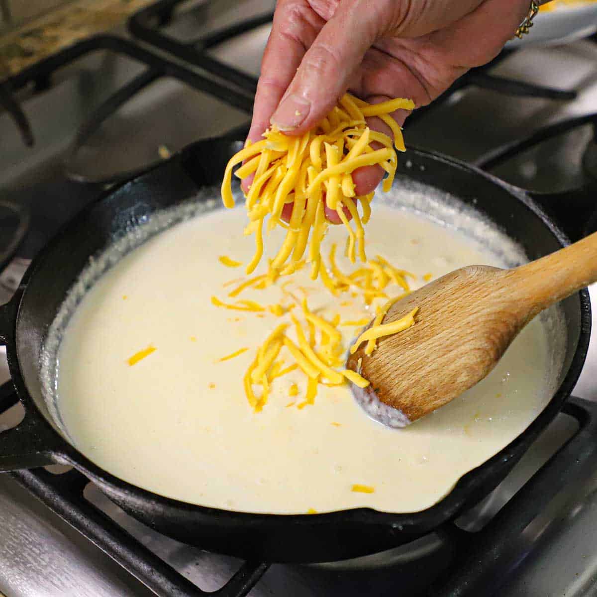 A person using his hand to sprinkle shredded yellow American cheese into a bechamel sauce in a black cast-iron skillet on a gas stove.