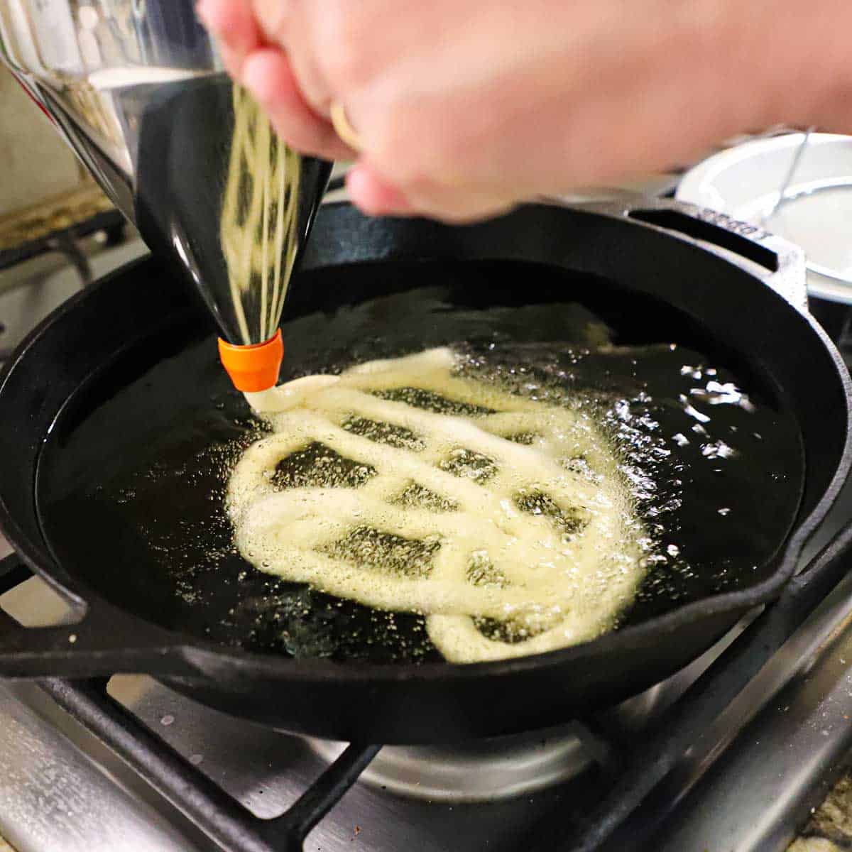 A person using a metal funnel batter dispenser to add circular rows of funnel cake batter into hot oil that is in a large cast-iron skillet on a gas stove.