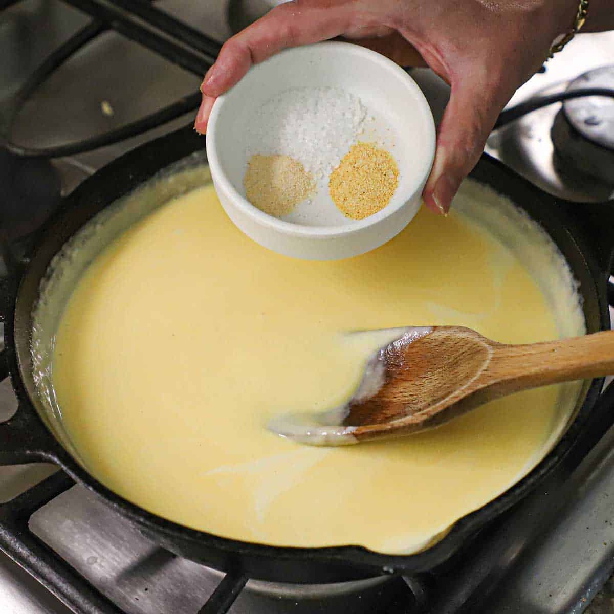 A person getting ready to transfer small amounts of garlic power, salt, and onion powder from a small bowl into a skillet filled with a simmering cheese sauce.