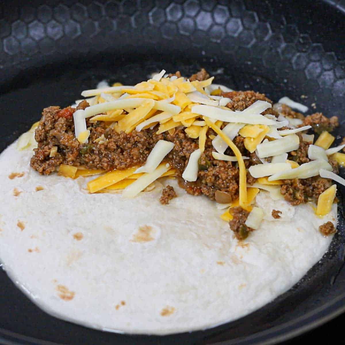 A flour tortilla in a non-stick skillet with taco meat and shredded cheese on one side of the tortilla.