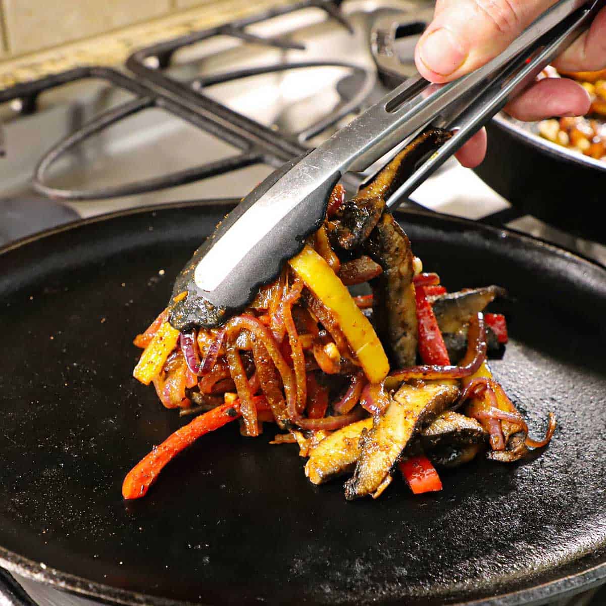 A person placing sautéed strips of mushrooms, bell peppers, and onions onto a fajita skillet that is being heated over a gas stove.