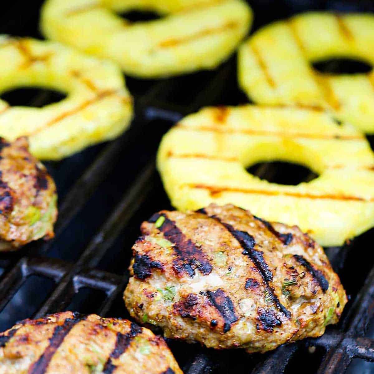 A cooked curry turkey burger resting on the grate of a gas grill next to rings of pineapple that are also being grilled.