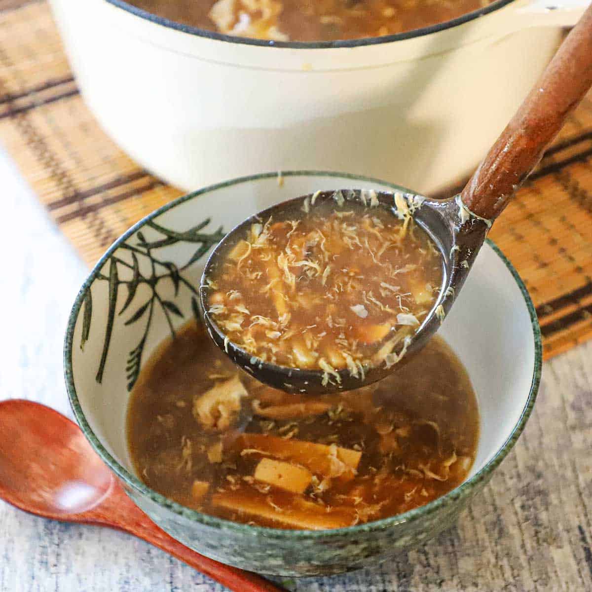 A person using a large wooden ladle to transfer homemade Chinese hot and sour soup from a large pot into a festive Asian soup bowl.