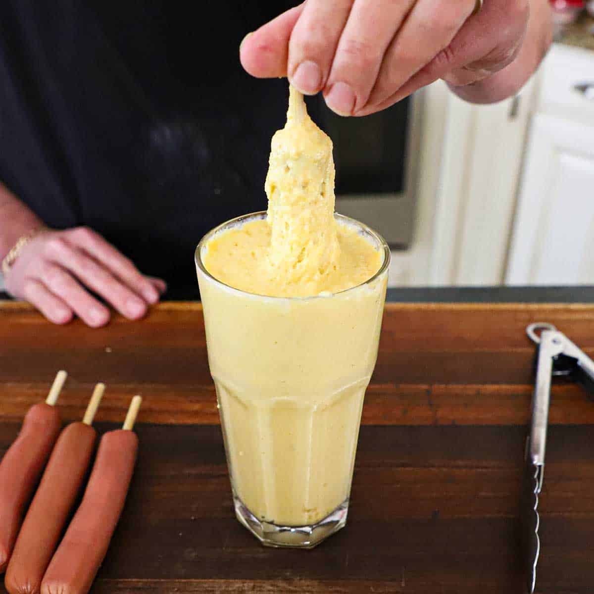 A person pulling a hot dog on a stick out of a glass that is filled with corn dog batter.