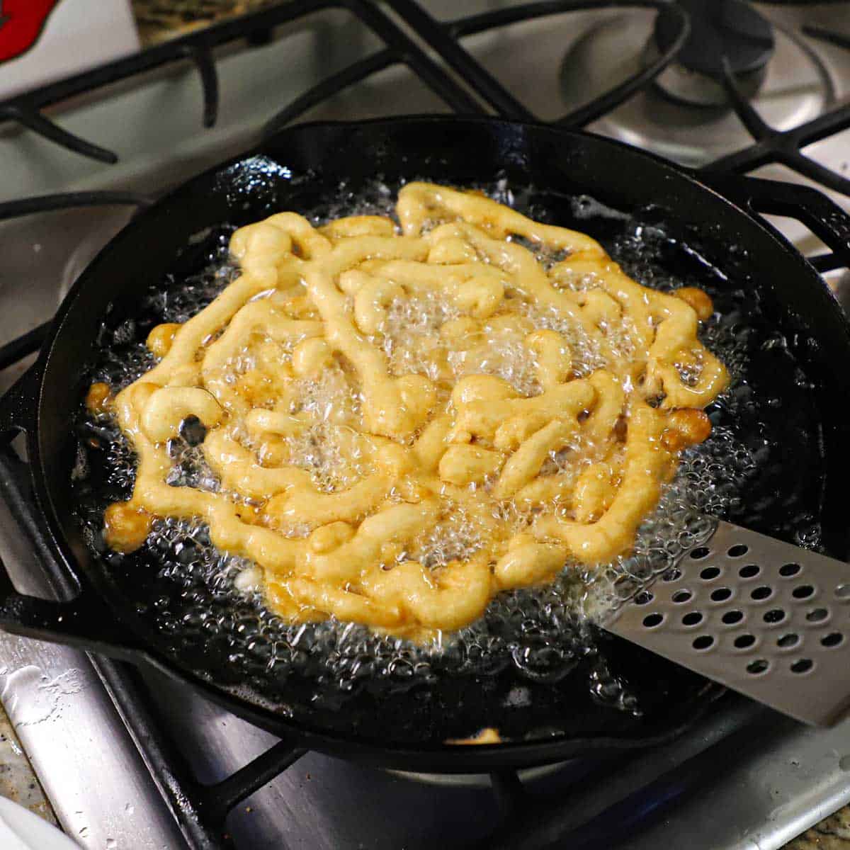 A person using a metal spatula to carefully flipped a partially cooked homemade funnel cake that is being fried in hot oil in a large cast-iron skillet on a gas stove.