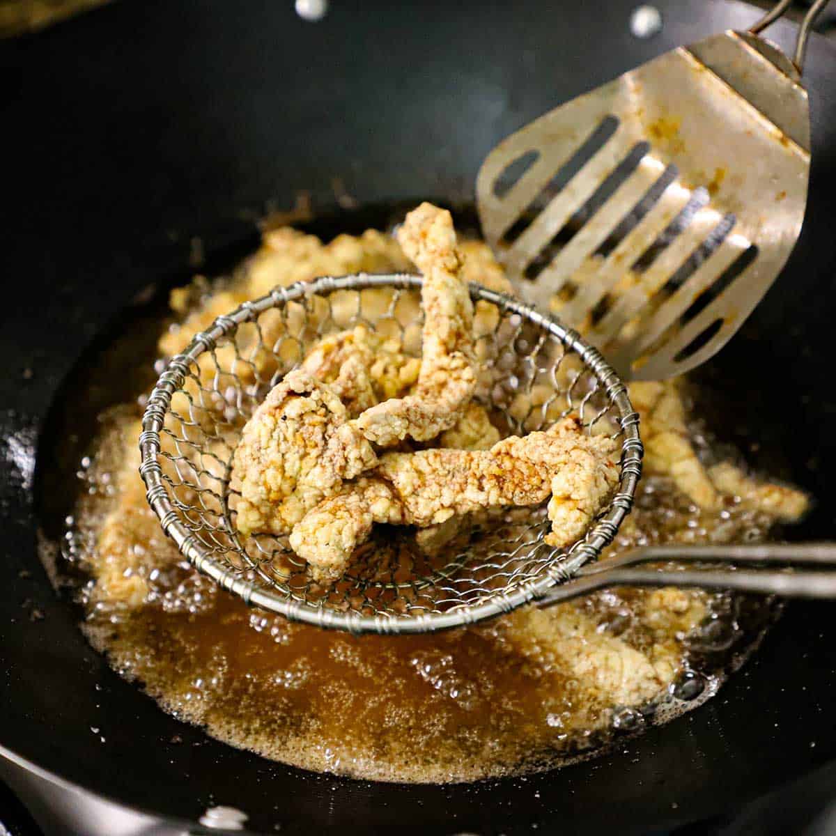 A person holding up a metal spider filled with crispy strips of breaded beef over a wok that also has crispy beef strips being deep-fried.