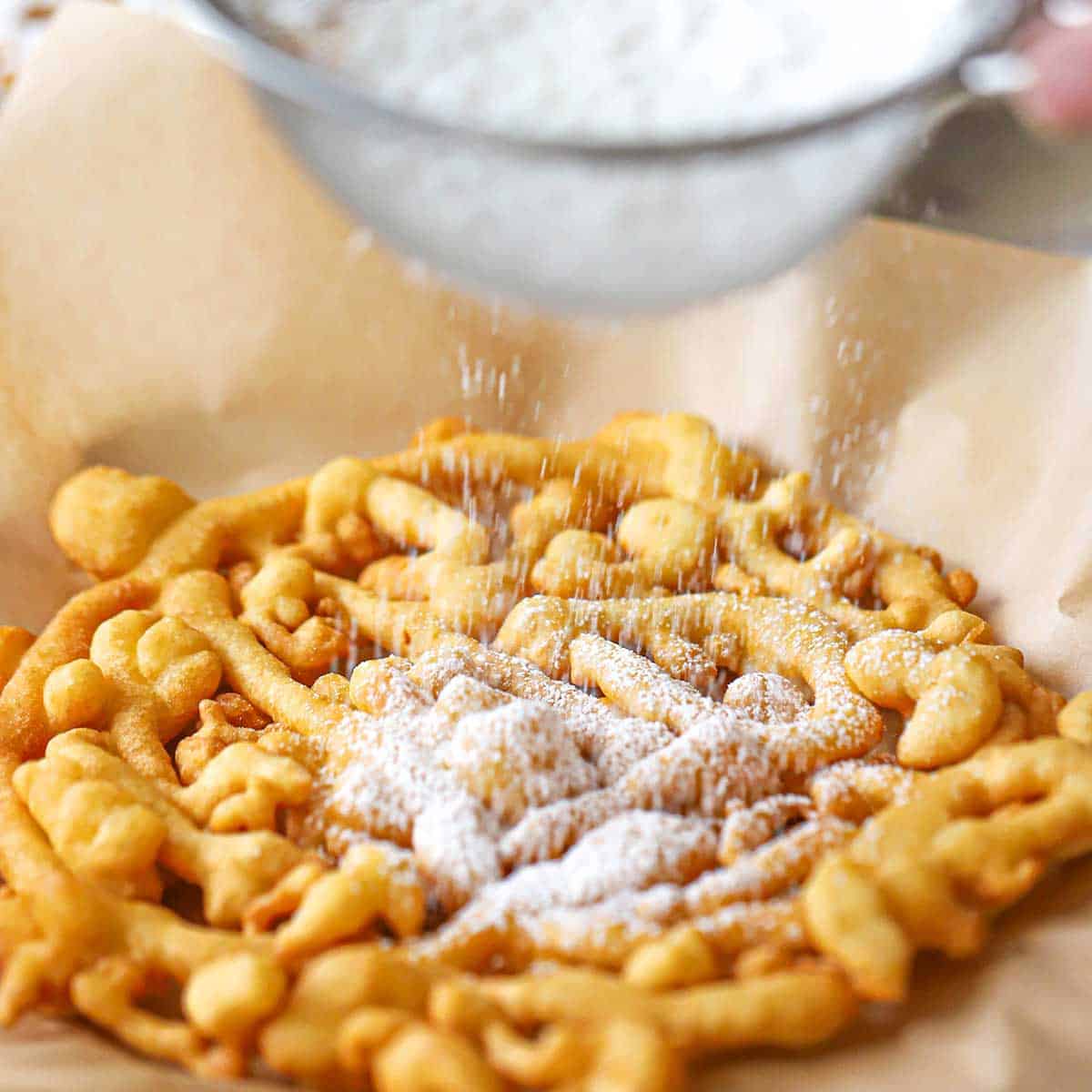 A person sifting powdered sugar from a sieve onto a freshly made homemade funnel cake that is resting on a brown piece paper.