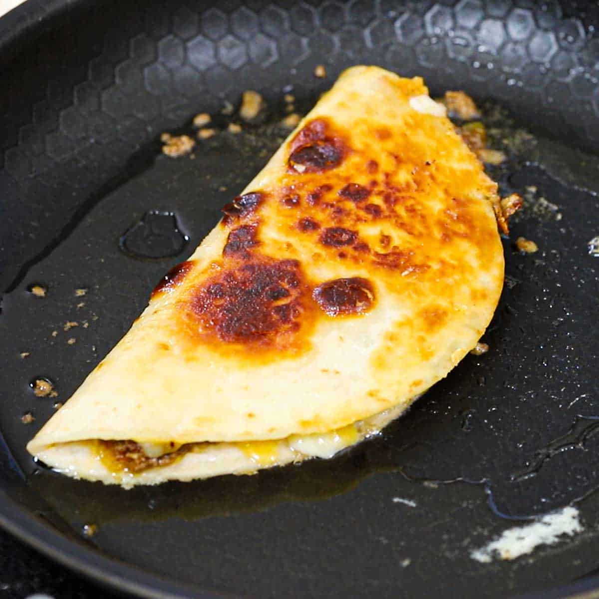 A freshly fried taco quesadilla sizzling in a non-stick skillet with oil simmering in the skillet.