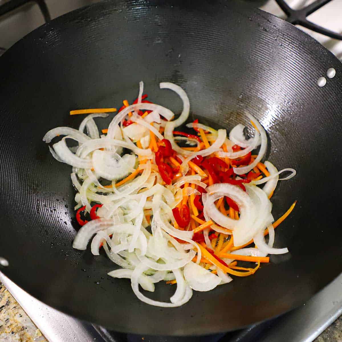 A large black wok sitting on a gas stove and is filled with thin strips of onion, julienned carrots, and slivers of a Fresno chili pepper.