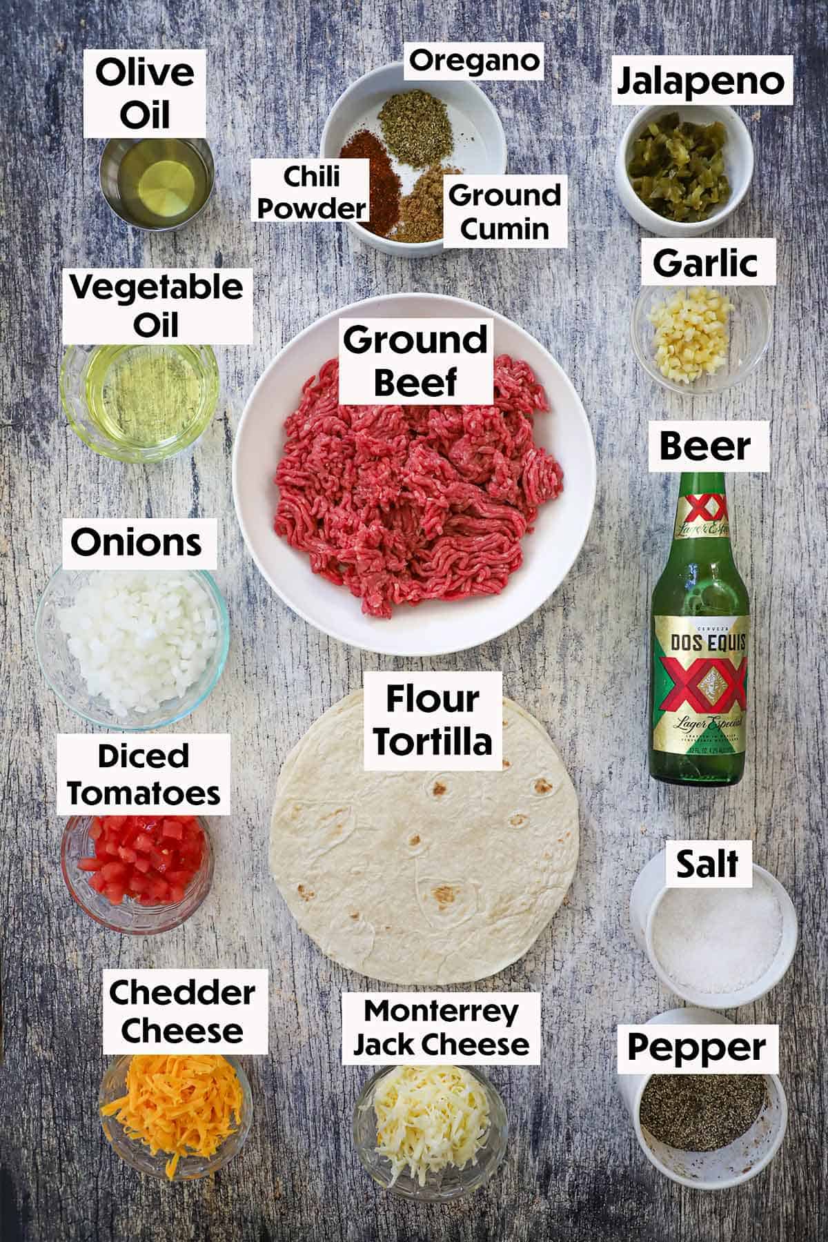 An arrangement of ingredients for taco quesadillas on a grey wooden background including ground beef, oil, beer, onions, seasonings, tomatoes, tortillas, and cheese.