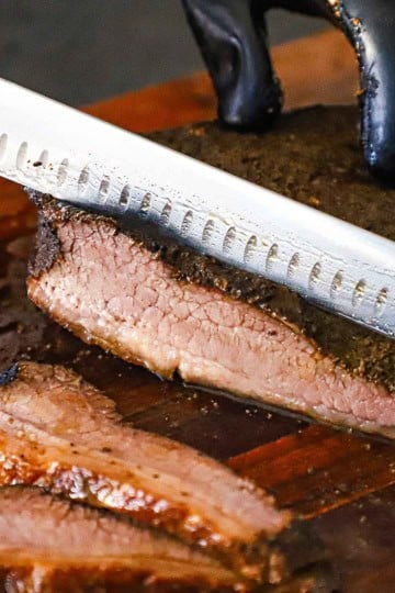 A person holding on a fully cooked Texas-Style Beef Brisket with one hand and using a large knife to slice it into juicy strips with the other hand.