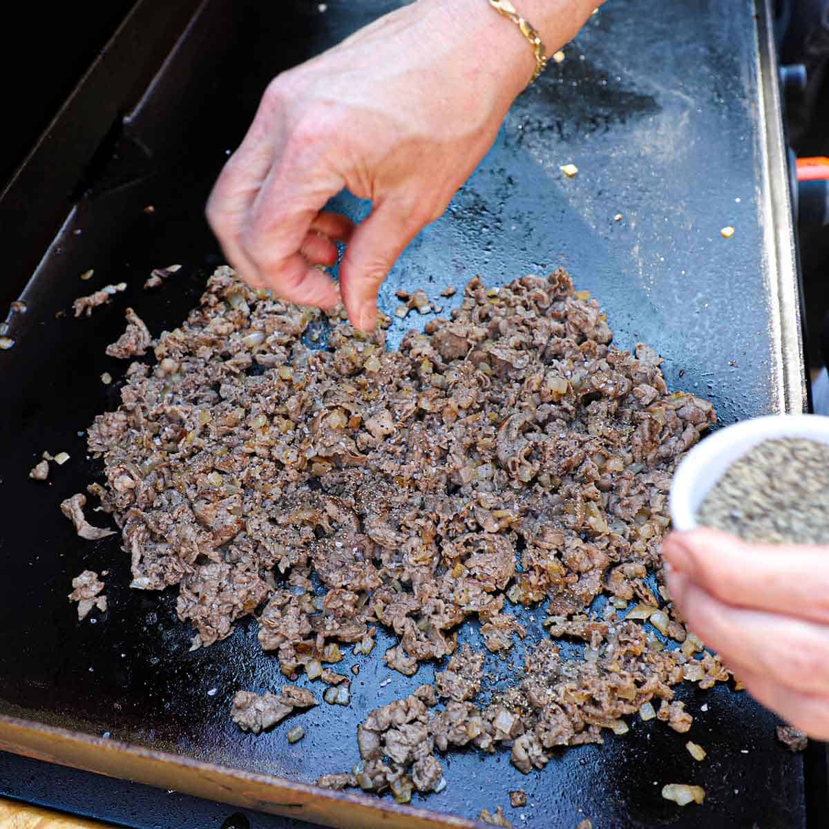 A person sprinkling ground pepper over chopped cooked ribeye and sautéed onions on a large outdoor griddle.