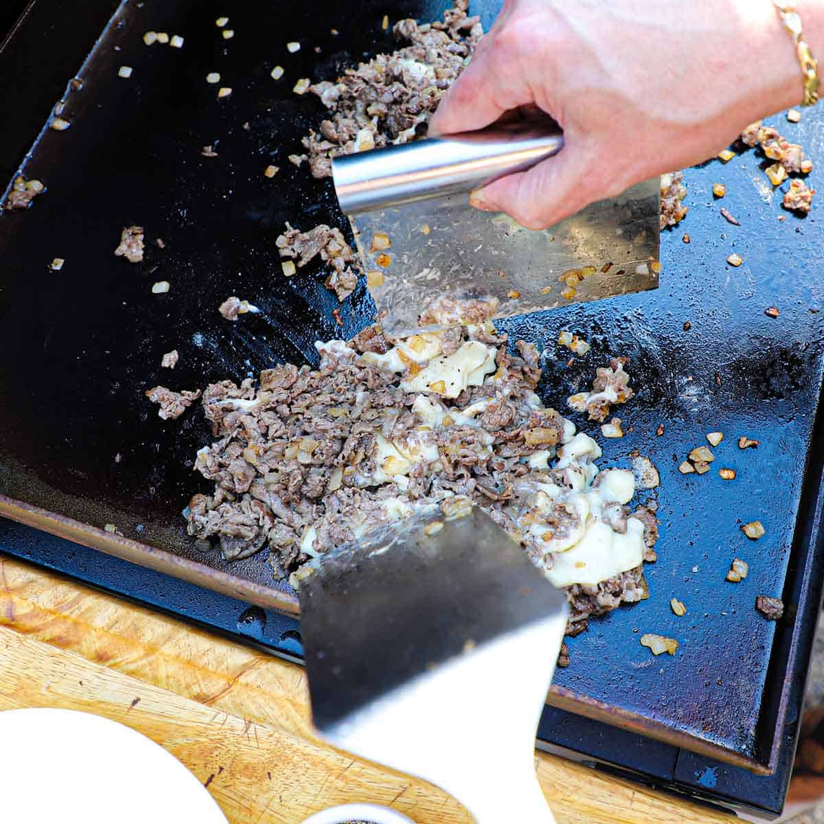 A person using a metal spatula and bench scraper to incorporate melty American white cheese into a pile of cooked thin strips of ribeye and sautéed onions on an outdoor griddle.
