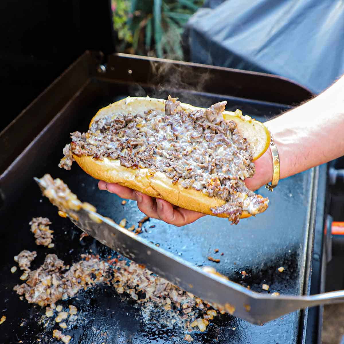 A person holding a homemade Philly cheesesteak in his right hand while holding the spatula that he used to transfer the meat into the bun with his other hand.